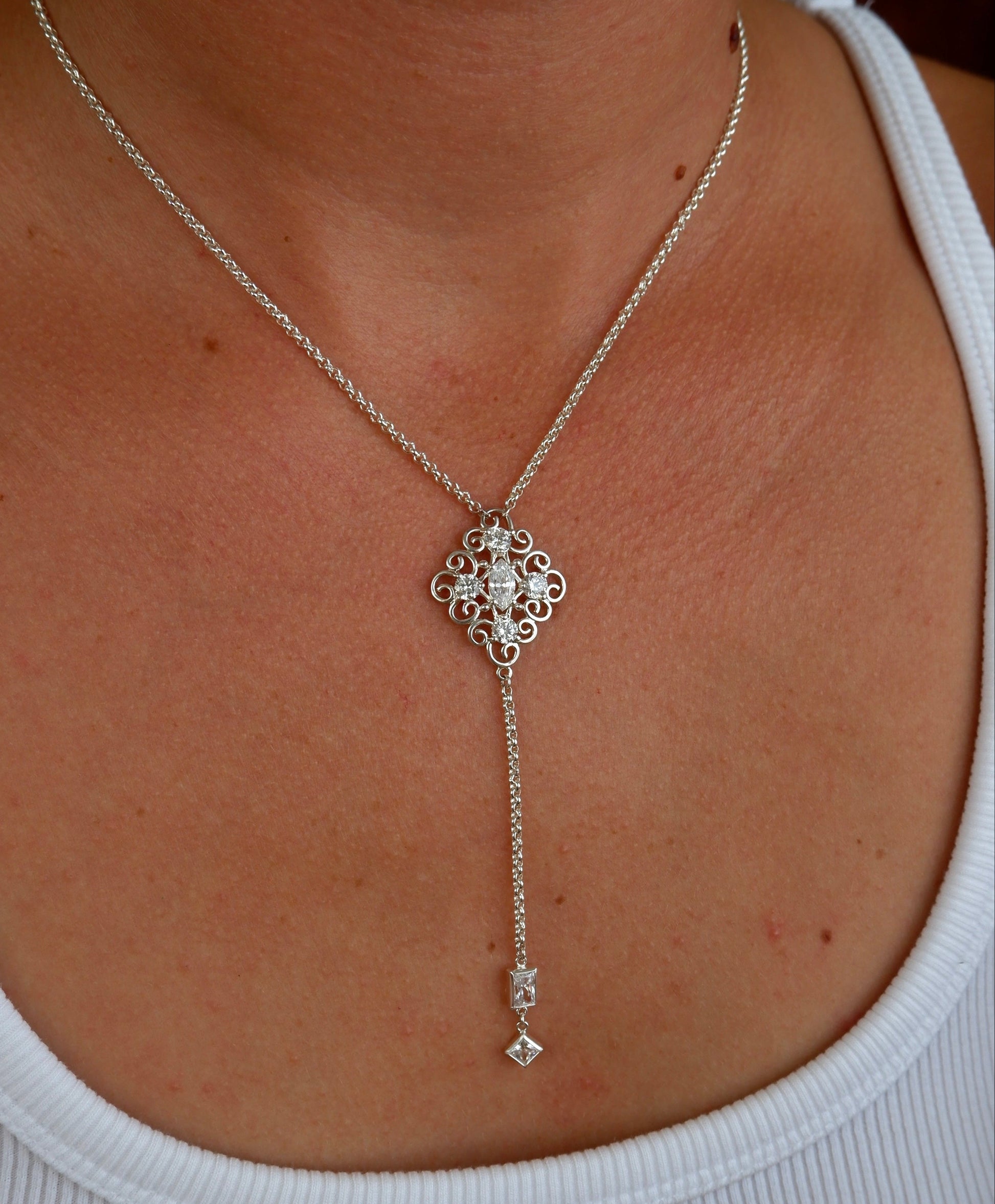 A silver lariat-style necklace with a decorative filigree pendant and a dangling charm, shown worn on a person's neck over a white ribbed tank top.