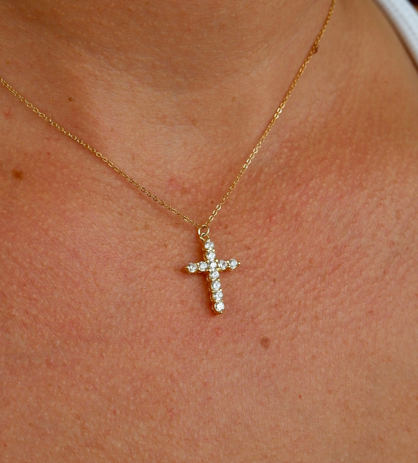 A close-up of a person's neck and chest wearing a delicate gold cross pendant adorned with small clear stones on a fine gold chain.