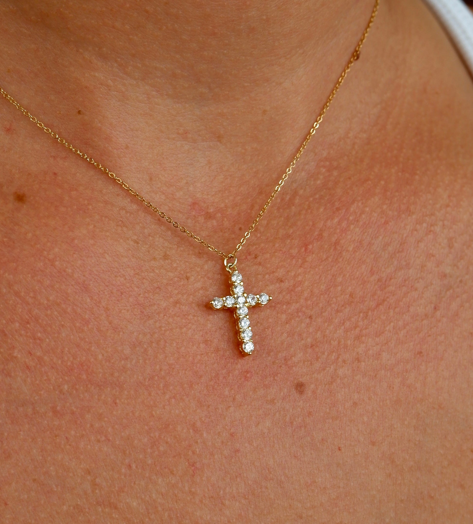 A close-up of a person's neck and chest wearing a delicate gold cross pendant adorned with small clear stones on a fine gold chain.