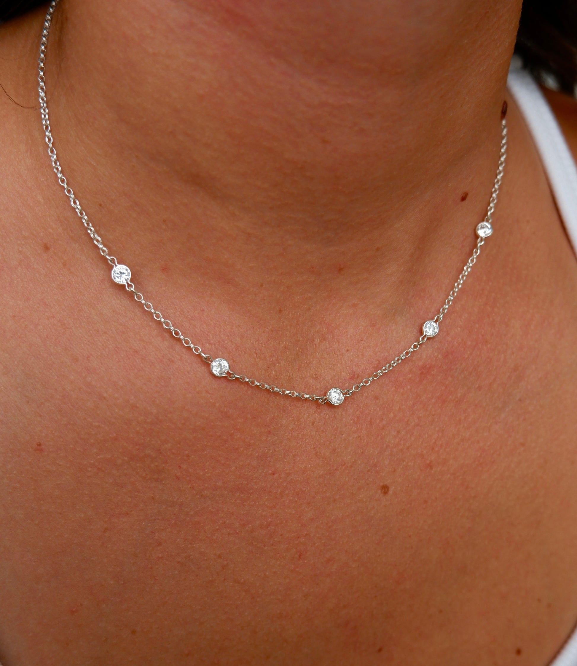 A close-up of a delicate silver chain necklace with five small round clear stones, worn on a person's neck over a white tank top.