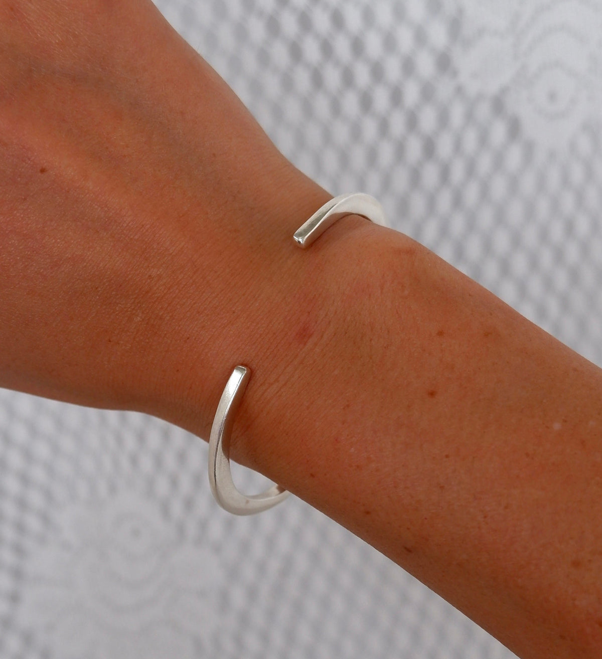A close-up of a person's wrist wearing a minimalist, open silver cuff bracelet against a textured light gray background.