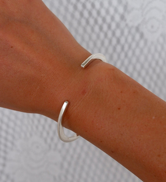 A close-up of a person's wrist wearing a minimalist, open silver cuff bracelet against a textured light gray background.