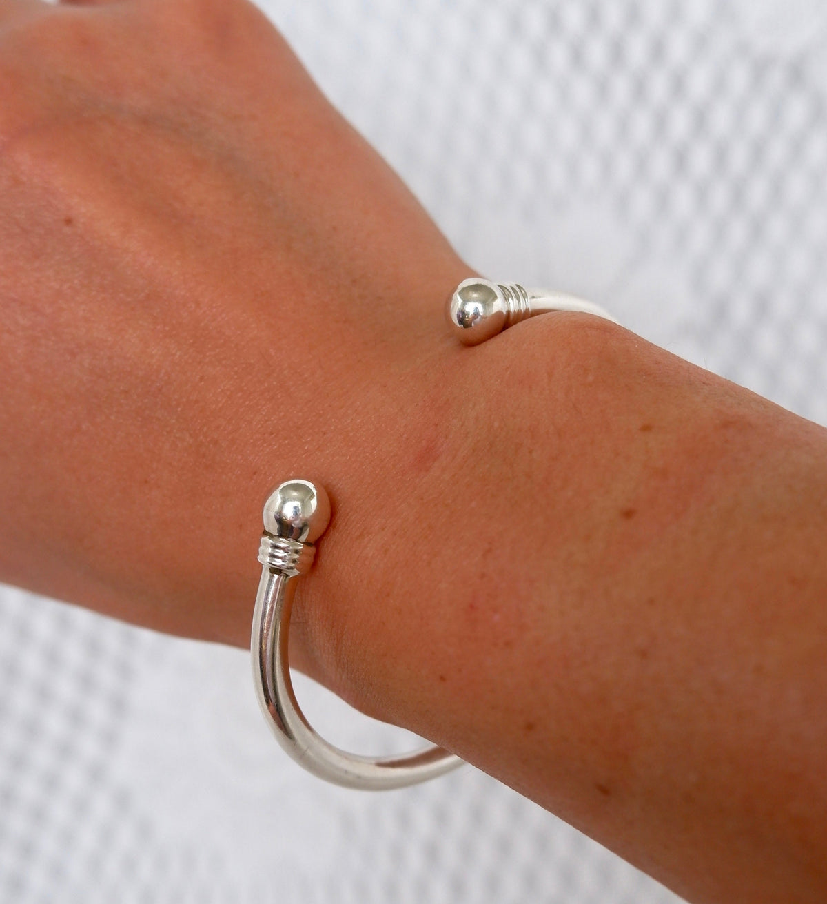 A close-up of a person's wrist wearing a polished silver cuff bracelet with decorative ball ends, set against a textured white background.