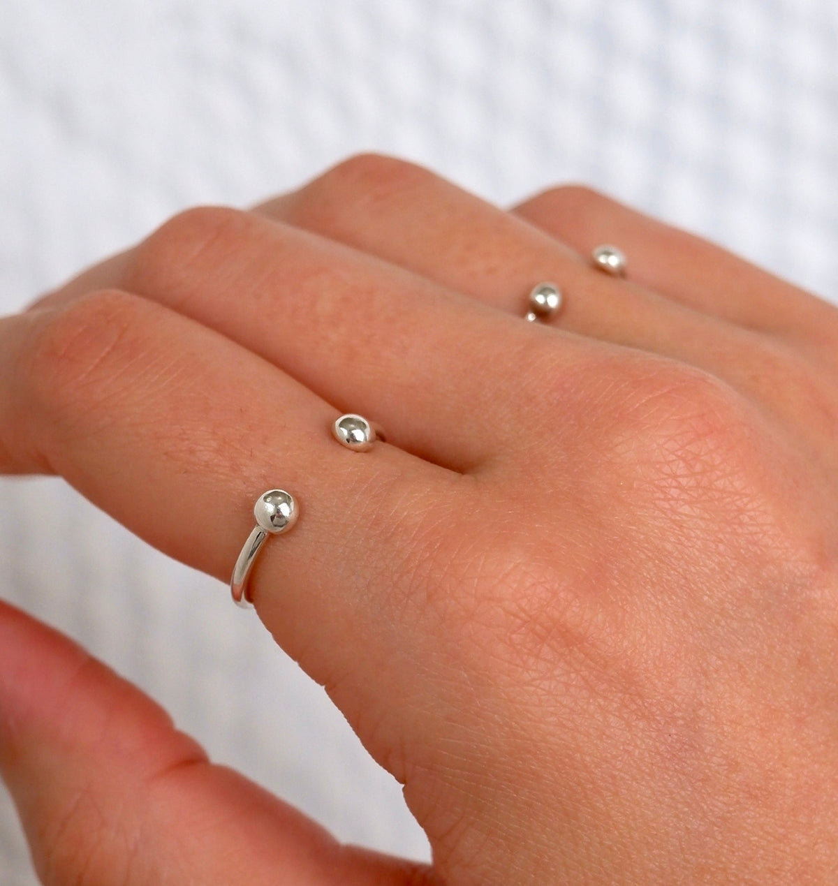 A close-up of a hand wearing three delicate silver rings, each featuring a small, polished bead on a thin band, set against a textured white background.