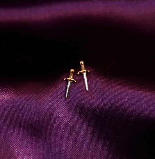Two gold sword-shaped earrings on a purple fabric background