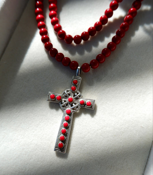 A red beaded necklace with a silver filigree cross pendant, decorated with small red stones, displayed on a white textured background.