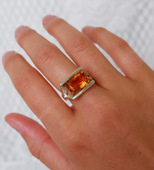 A silver ring featuring a large, faceted orange gemstone, shown worn on a person's finger against a textured white background.