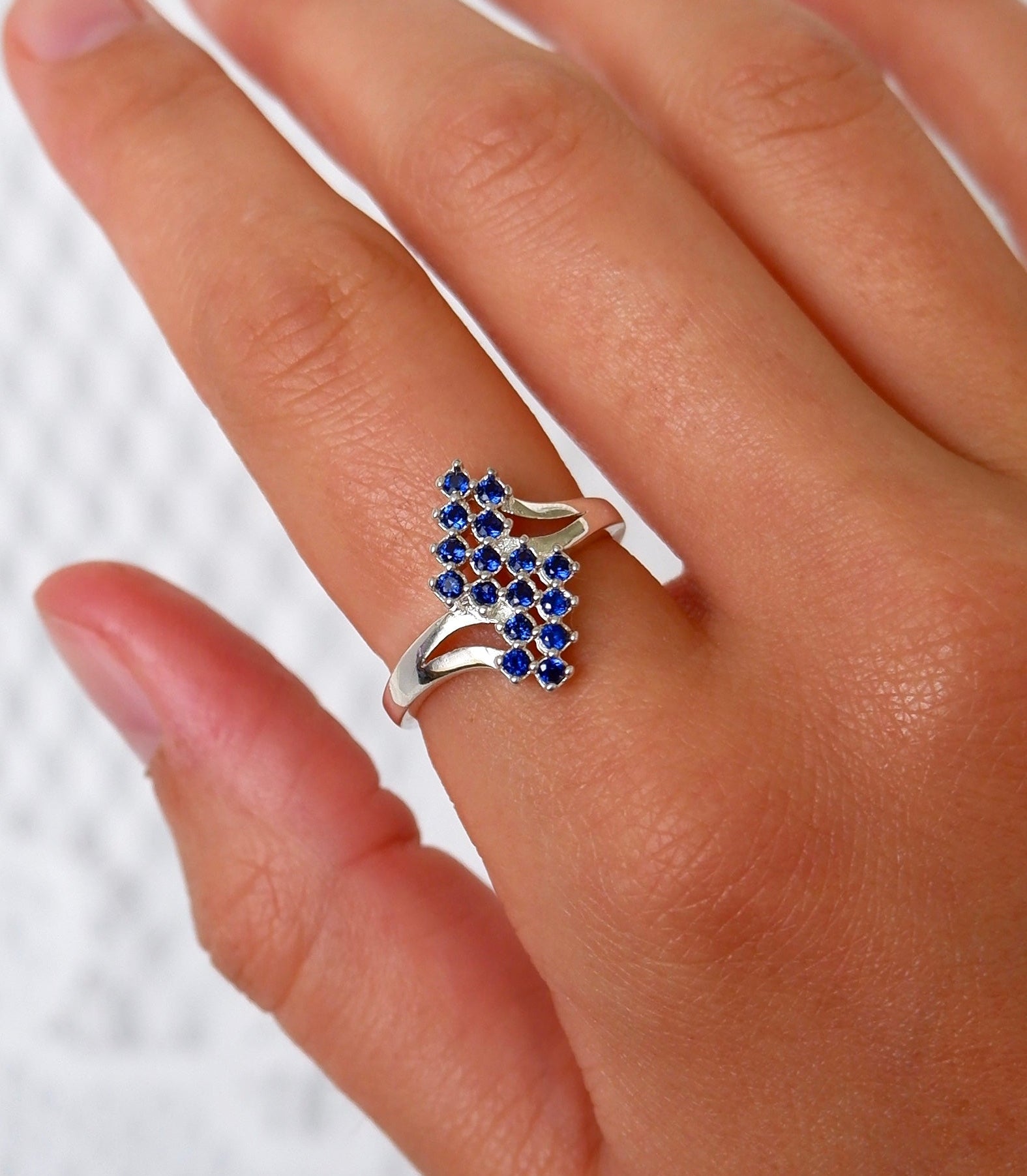 A close-up of a hand wearing a silver ring with a cluster of small round blue gemstones on the ring finger, set against a light textured background.