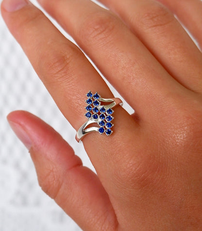 A close-up of a hand wearing a silver ring with a cluster of small round blue gemstones on the ring finger, set against a light textured background.