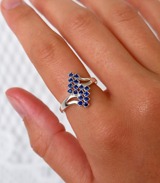 A close-up of a hand wearing a silver ring with a cluster of small round blue gemstones on the ring finger, set against a light textured background.