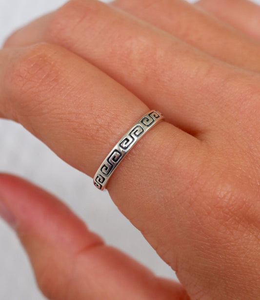 A close-up of a hand wearing a thin silver band ring with a Greek key pattern engraved on the side.
