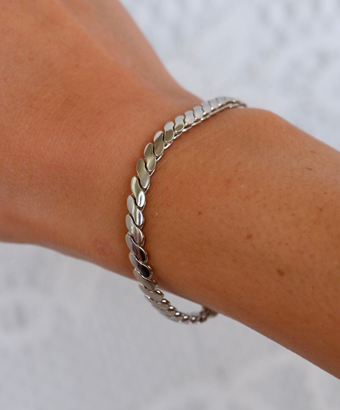 A close-up of a person's wrist wearing a silver leaf-link chain bracelet against a textured white background.
