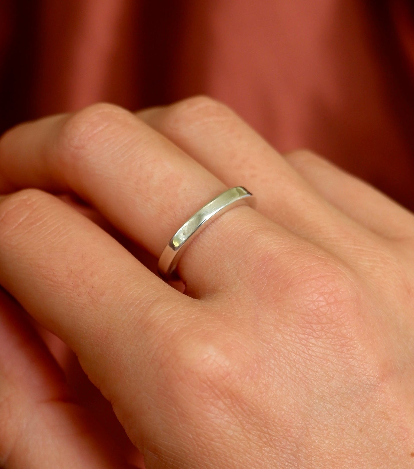 A close-up of a hand wearing a simple polished silver band ring against a warm rust-colored fabric background.